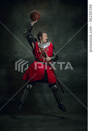 Portrait of medieval warrior or knight with dirty wounded face playing with basketball ball isolated over dark background. Comparison of eras, history Portrait of medieval warrior or knight with dirty wounded face playing with basketball ball isolated over dark background. Comparison of eras, history 86280166