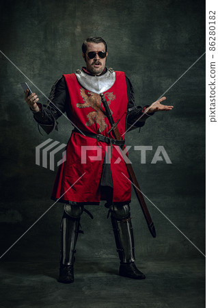 Surprised young man in image of medieval warrior or knight with dirty wounded face using phone isolated over dark background. Comparison of eras Surprised young man in image of medieval warrior or knight with dirty wounded face using phone isolated over dark background. Comparison of eras 86280182