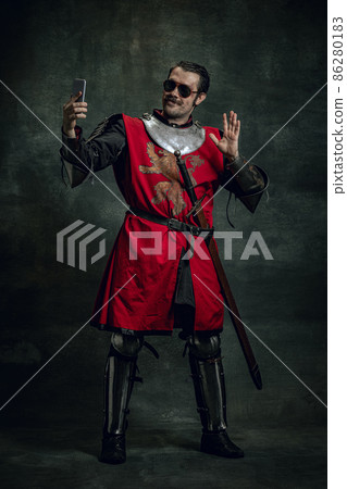 Smiling young man in image of medieval warrior or knight with dirty wounded face using phone isolated over dark background. Comparison of eras Smiling young man in image of medieval warrior or knight with dirty wounded face using phone isolated over dark background. Comparison of eras 86280183