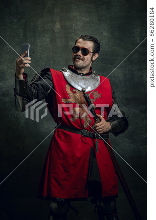 Smiling young man in image of medieval warrior or knight with dirty wounded face using phone isolated over dark background. Comparison of eras Smiling young man in image of medieval warrior or knight with dirty wounded face using phone isolated over dark background. Comparison of eras 86280184
