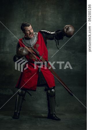 Portrait of medieval warrior or knight with dirty wounded face in boxing gloves isolated over dark background. Comparison of eras, history 86280186