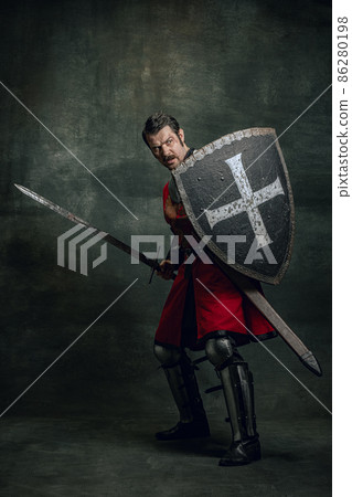 Portrait of medieval warrior or knight with dirty wounded face holding shield and sword isolated over dark background. Comparison of eras, history 86280198