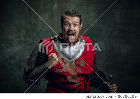 Comic portrait of funny medieval warrior or knight with dirty wounded face holding sword isolated over dark background. Comparison of eras 86280199