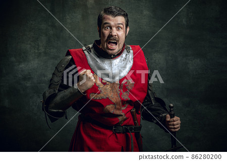 Half-length portrait of brutal seriuos man, medieval warrior or knight with dirty wounded face holding sword isolated over dark background. Comparison of eras 86280200