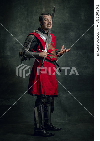 Comic portrait of funny medieval warrior or knight with dirty wounded face holding sword isolated over dark background. Comparison of eras 86280201