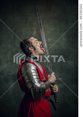 Comic portrait of funny medieval warrior or knight with dirty wounded face holding sword isolated over dark background. Comparison of eras 86280202