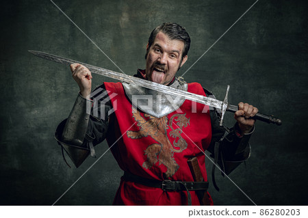 Comic portrait of funny medieval warrior or knight with dirty wounded face holding sword isolated over dark background. Comparison of eras 86280203