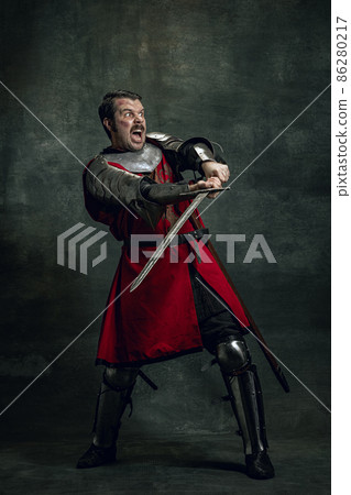 Dynamic portrait of seriuos man, medieval warrior or knight with dirty wounded face holding sword isolated over dark background. Comparison of eras Dynamic portrait of seriuos man, medieval warrior or knight with dirty wounded face holding sword isolated over dark background. Comparison of eras 86280217