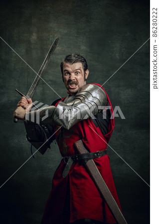 Dynamic portrait of seriuos man, medieval warrior or knight with dirty wounded face holding sword isolated over dark background. Comparison of eras Dynamic portrait of seriuos man, medieval warrior or knight with dirty wounded face holding sword isolated over dark background. Comparison of eras 86280222