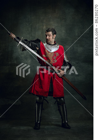 Comic portrait of funny medieval warrior or knight with dirty wounded face holding sword isolated over dark background. Comparison of eras Comic portrait of funny medieval warrior or knight with dirty wounded face holding sword isolated over dark background. Comparison of eras 86280270
