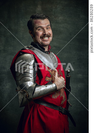 Portrait of smiling man in image of medieval warrior with dirty wounded face wearing equipment isolated over dark background. Comparison of eras Portrait of smiling man in image of medieval warrior with dirty wounded face wearing equipment isolated over dark background. Comparison of eras 86280289