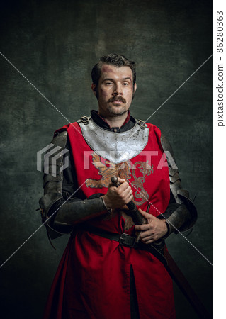 Vintage style portrait of brutal seriuos man, medieval warrior or knight with dirty wounded face holding sword isolated over dark background. Comparison of eras 86280363