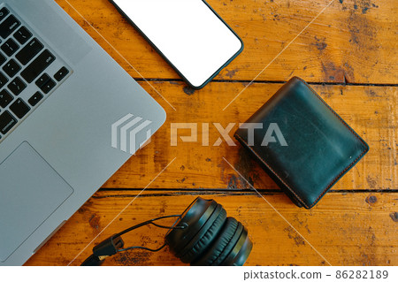 Top view of laptop, headphones, smartphone, and wallet on wooden table. Freelancer's work surface. Blank mobile phone screen to insert some text. Modern devices for business jobs and entertainment. 86282189