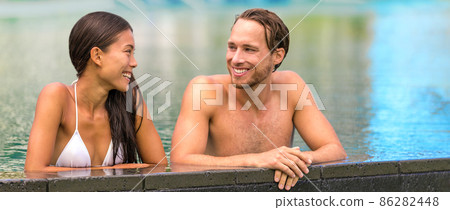 Couple relaxing at luxury pool resort banner panoramic. Young people Asian woman, Caucasian man enjoying summer holiday in travel vacation destination talking and laughing together. 86282448