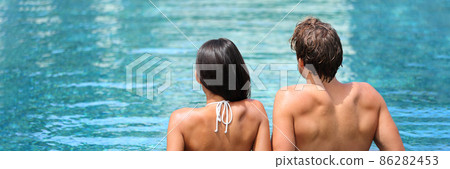 Luxury spa couple relaxing at swimming pool resort panoramic. Adults only hotel travel vacation honeymoon getaway. Woman and man relaxation concept. Two people from behind enjoying summer holidays. 86282453