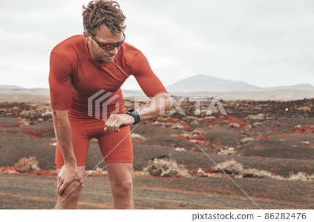 Tired runner man checking wearable technology sports smartwatch during outdoor training. Sport athlete looking at watch during workout on desert trail. Man wearing sunglasses, compression outfit. 86282476