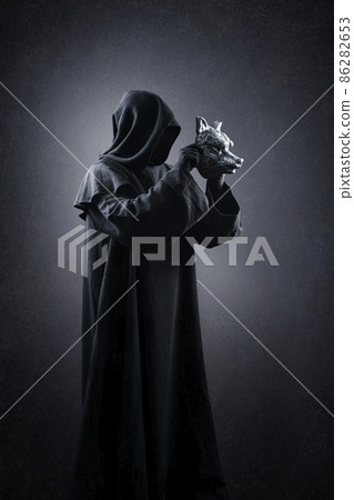 Creepy figure in hooded cloak with wolf mask in hands over dark misty background 86282653