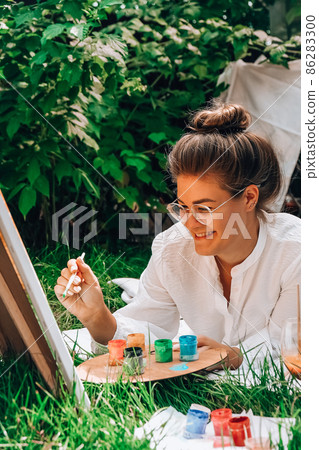 Artist painting on the easel outdoors in the garden. Open air outdoor art workshop. Draw on the canvas with brush and palette sitting on the grass during a picnic in a park 86283300