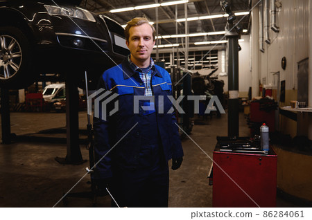 A technician, auto mechanic, car engineer standing against the background of a lifted automobile with wrench in his hand, working in repair shop or car service garage 86284061
