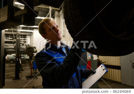 A young competent auto mechanic inspects the car raised on the elevator for damage. Concept of technical maintenance and car service A young competent auto mechanic inspects the car raised on the elevator for damage. Concept of technical maintenance and car service 86284069