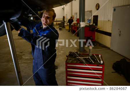 Handsome Caucasian young man, car engineer, technician, auto mechanic repairing a lifted modern automobile on a hoist in the repair shop garage. Automobile maintenance and auto service concept Handsome Caucasian young man, car engineer, technician, auto mechanic repairing a lifted modern automobile on a hoist in the repair shop garage. Automobile maintenance and auto service concept 86284087