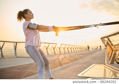 Young sportswoman practicing body weight training outdoors early in the morning at dawn. Female athlete exercising with suspension straps on the city bridge at sunrise. Sport, active lifestyle concept 86284202