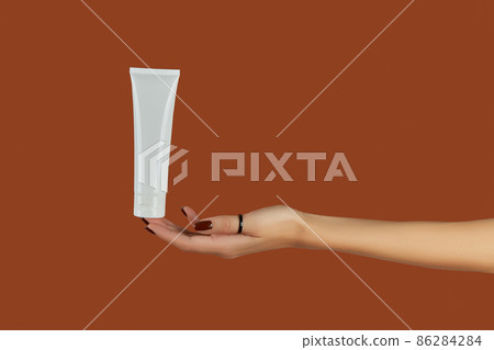 Womans hand holding white tube on brown background. Self care concept. Womans hand holding white tube on brown background. Self care concept. 86284284