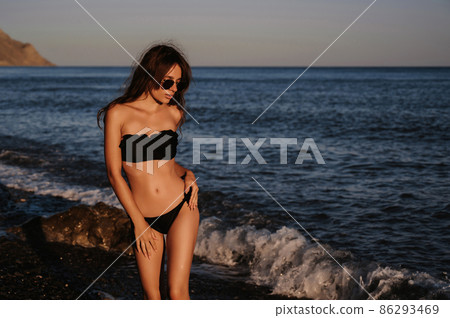 young girl with a sexy body in a black swimsuit and sunglasses enjoys relaxing on the beach by the sea 86293469