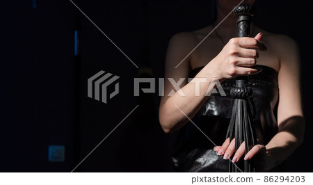 BDSM concept. A woman in a leather dress holds a black whip with a long fringe. Role-playing games for adults. Close-up of sex toys for domination. 86294203