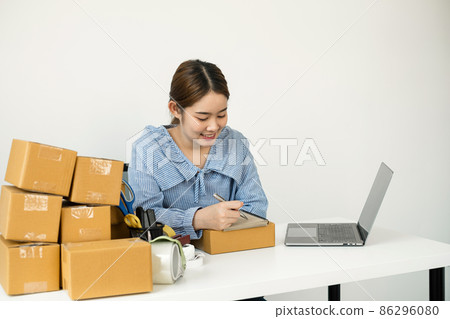 Young Asian woman business owner with many parcel boxes on the table happy online sales job, use the laptop, get an order from customers, take notes, and make arrangements for delivery by post 86296080