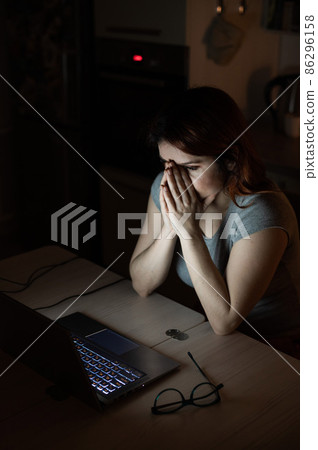 woman works overtime. A student studying at a laptop at night in the dark. The girl is worried about problems at work and holds her hands on her face. woman works overtime. A student studying at a laptop at night in the dark. The girl is worried about problems at work and holds her hands on her face. 86296158