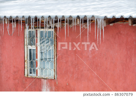 Ice icicles hang from the roof above the barred window of an old house with a red wall. 86307079