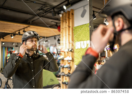 Man examining bicycle helmets in sport shop looking at herself in mirror. Trying new sports helmet in bike shop. Male putting on cycle helmet in shop. Customer in bicycle store trying on bike helmet Man examining bicycle helmets in sport shop looking at herself in mirror. Trying new sports helmet in bike shop. Male putting on cycle helmet in shop. Customer in bicycle store trying on bike helmet 86307458