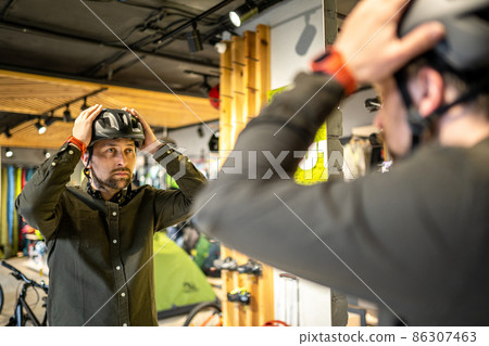 Caucasian man trying on black bicycle helmet near mirror in sporting goods store. Male buyer chooses safety helmet for cycling. Shopping in bicycle store. Person adjusts his sports helmet strap 86307463