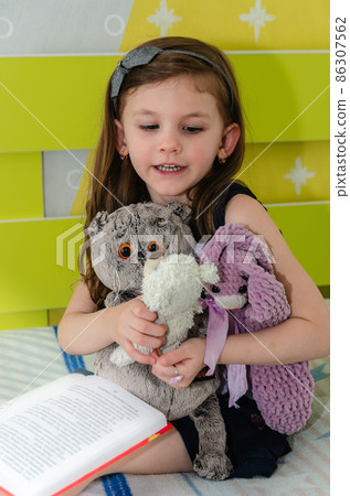 A little girl is reading a book of fairy tales to her toys in her room on the bed. A little girl is reading a book of fairy tales to her toys in her room on the bed. 86307562
