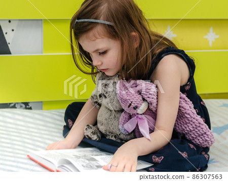 A little girl is reading a book of fairy tales to her toys in her room on the bed. A little girl is reading a book of fairy tales to her toys in her room on the bed. 86307563
