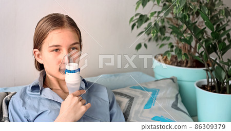 Teenage girl makes inhalation with a nebulizer equipment. Sick child holding inhalator in hand and breathes through an inhaler at home 86309379