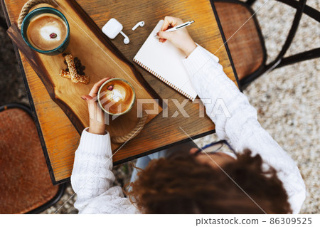 Girl hands with cup of coffee and blank diary with pencil on a wooden table. Girl hands with cup of coffee and blank diary with pencil on a wooden table. 86309525