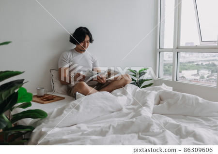 Asian man reading book on the bed with breads and coffee in the apartment. Asian man reading book on the bed with breads and coffee in the apartment. 86309606