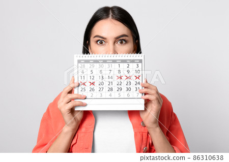 Pensive young woman holding calendar with marked menstrual cycle days on beige background 86310638