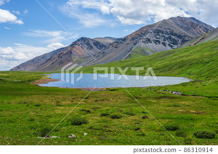 Colorful summer landscape with beautiful lake in sunlit green mountain valley among rocks and high mountain ridge under blue sky. Awesome scenery with alpine lake among greenery in sunlight. 86310914