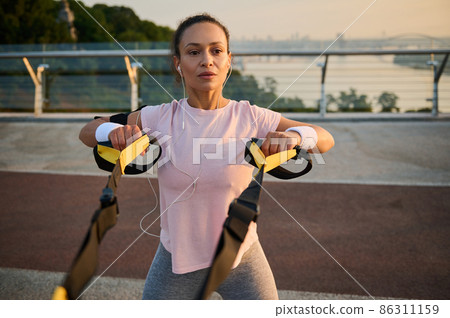 Determined female athlete doing bodyweight training with suspension straps against a bridge and river background. Sportswoman exercising outdoor at dawn. Sport, fitness, active lifestyle concept 86311159