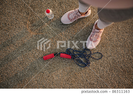 High angle view of female athlete legs wearing pink sneakers and standing on an asphalt treadmill next to a lying down skipping rope and water bottle High angle view of female athlete legs wearing pink sneakers and standing on an asphalt treadmill next to a lying down skipping rope and water bottle 86311163