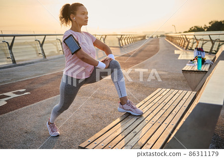 Curly dark-haired sporty woman in active wear with smartphone holder stretching and flexing her legs muscles on a wooden bench while working out outdoor at sunrise Curly dark-haired sporty woman in active wear with smartphone holder stretching and flexing her legs muscles on a wooden bench while working out outdoor at sunrise 86311179