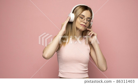 Beautiful attractive young blond woman wearing pink t-shirt and glasses in white headphones listening music on pink background in studio. Lifestyle. Beautiful attractive young blond woman wearing pink t-shirt and glasses in white headphones listening music on pink background in studio. Lifestyle. 86311403