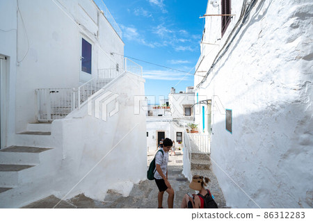 Ostuni, Puglia, Italy. August 2021. A young Caucasian with his mother are getting some fresh air in the shade in one of the white alleys of the historic center of the village. Vacation concept. 86312283