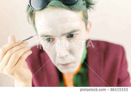 Mim is preparing for performance. Cosplayer guy paints his face with brush. Actor with white makeup and green hair in burgundy suit with yellow tie. 86315333