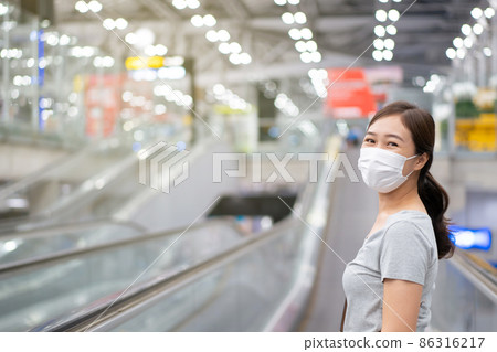Beautiful Asian young woman with protective face mask looking and smiling to the camera while walking in the airport. Travel and tourist concept during the coronavirus or COVID-19 pandemic. Beautiful Asian young woman with protective face mask looking and smiling to the camera while walking in the airport. Travel and tourist concept during the coronavirus or COVID-19 pandemic. 86316217