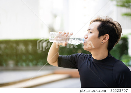 Active Asian sportsman relaxing and drinking a water in bottle after outdoor running or workout. Asian man drink a water during the break from exercise. A healthy man trying hard for cardio workout. Active Asian sportsman relaxing and drinking a water in bottle after outdoor running or workout. Asian man drink a water during the break from exercise. A healthy man trying hard for cardio workout. 86323707