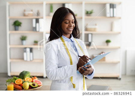Cheerful black nutritionist with clipboard prescribing healthy diet or creating plant based meal plan for patient 86326941